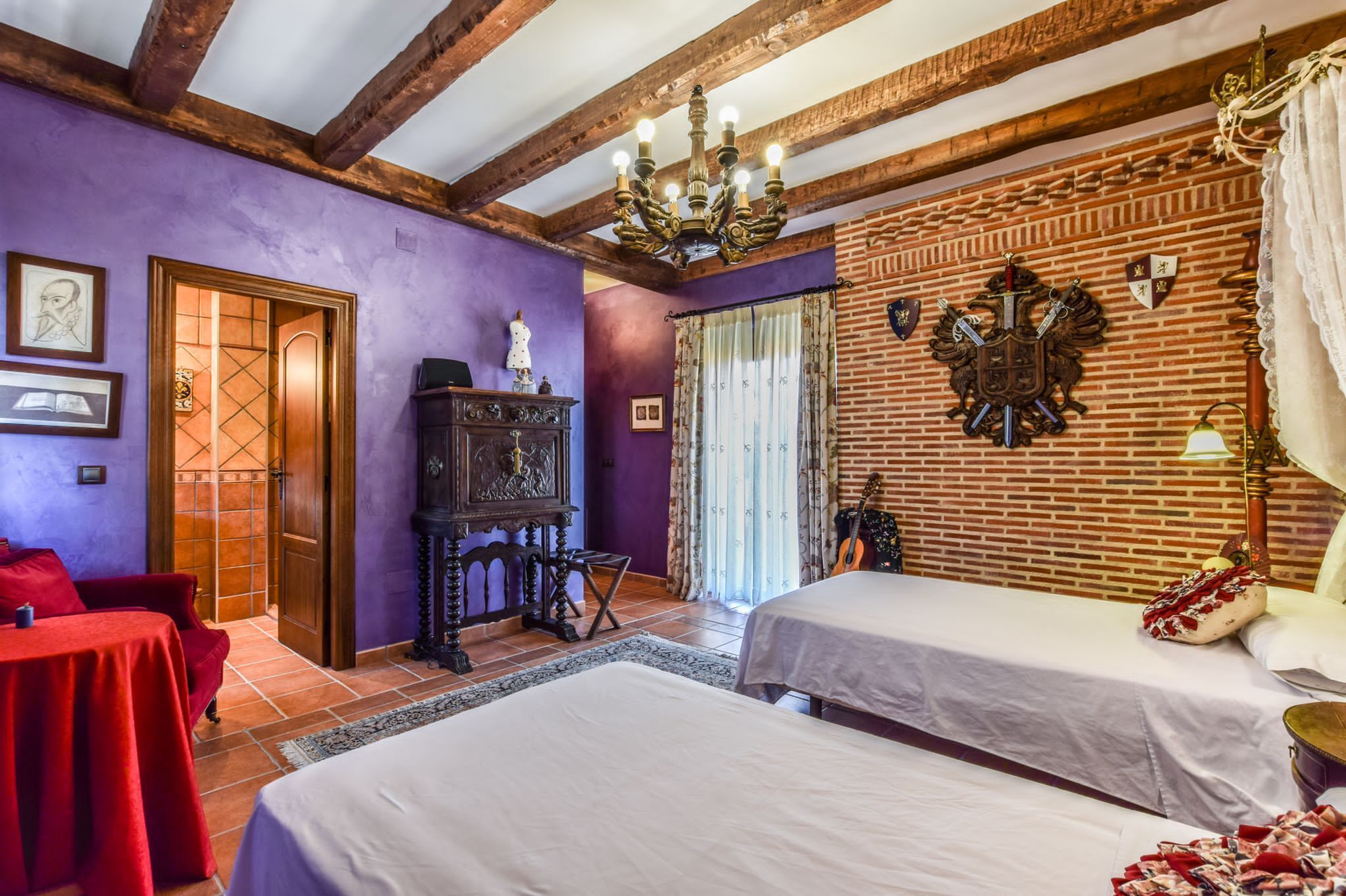 A room with two beds, a wooden ceiling, a brick wall, and a chandelier.