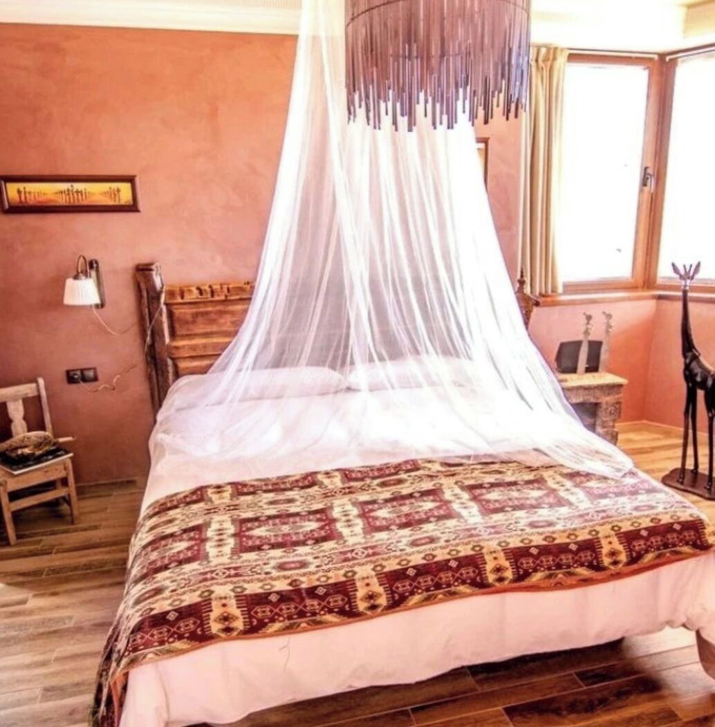 Un dormitorio con cama con dosel, dosel, colcha estampada, mesita de noche de madera, lámpara, pequeña mesa con una figurita de jirafa y una ventana con cortinas.
