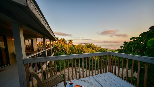 A wooden deck with a table and chairs, overlooking a beach and palm trees.