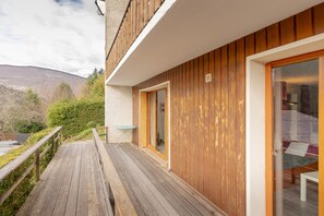 Una terraza de madera que conduce a un edificio con puerta de cristal, una mesita y vistas a la montaña.