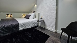 A bedroom with a sloped ceiling, a skylight, a bed with a dark patterned bedspread, a side table with a lamp, and a chair.