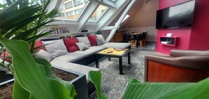 Ett modernt vardagsrum med högt tak, ett takfönster, en soffgrupp, ett soffbord och en tv som är monterad på en rosa vägg.