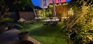 Ein Terrassenbereich im Freien mit Tischen und Stühlen, umgeben von Grünpflanzen und Lichterketten.