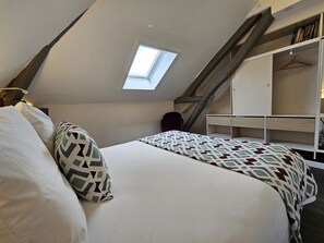 Une chambre avec un plafond en pente, un puits de lumière, un lit agrémenté d’oreillers à motifs, une chaise rouge et une armoire blanche avec des étagères.
