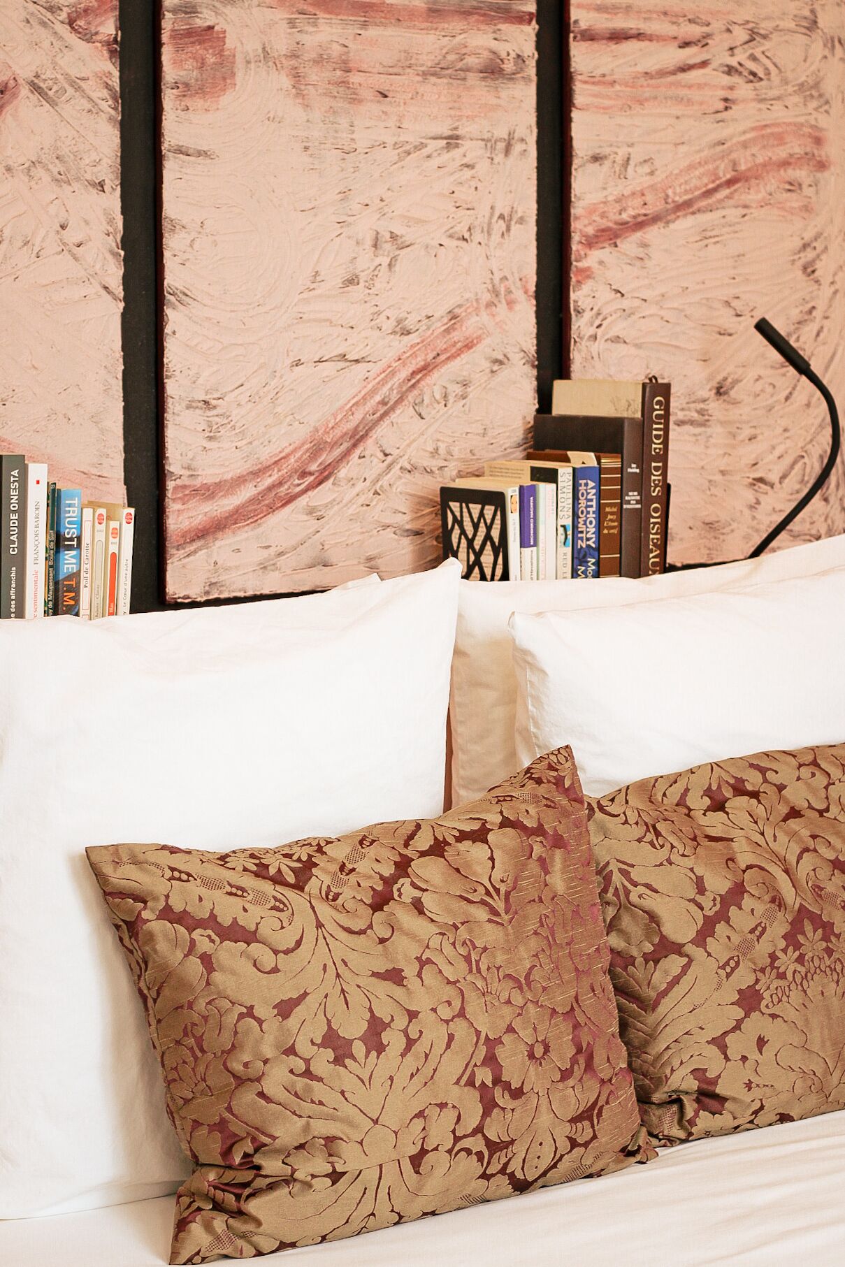Ein Bett mit weißer Bettwäsche und zwei gemusterten Kissen, ein Bücherregal mit Büchern sowie eine Wand mit strukturierter Gestaltung.