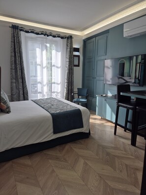 A bedroom with a bed, a desk, and a chair. There is a window with curtains, a flat-screen TV, and a door.