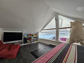 Une pièce avec un plafond en pente, une grande fenêtre, une télévision, un canapé et un ours en peluche.