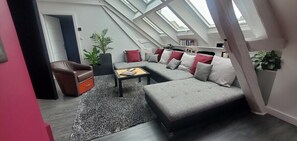 A modern loft-style living room with a grey sectional sofa, a brown leather armchair, and a wooden coffee table. There is a skylight above the sofa and a large potted plant to the left. - Apartment-Luxury-Private Bathroom-Garden view-Rdc A (Troyes)
