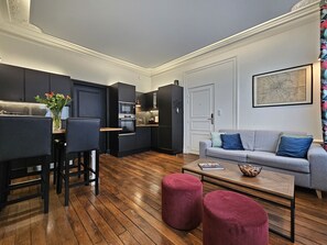 Un salon moderne avec un canapé gris, des poufs rouges, une table basse et un coin cuisine doté de meubles et d’appareils électroménagers aux couleurs sombres.