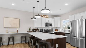 Une cuisine moderne dotée d’un îlot central, de tabourets de bar, d’appareils électroménagers en acier inoxydable et de luminaires suspendus.