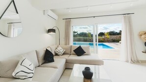 Een moderne woonkamer met een beige bank, een glazen salontafel en uitzicht op een zwembad door grote glazen deuren.