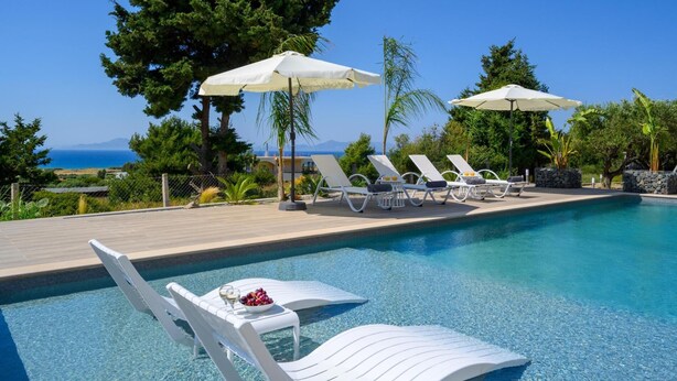 A poolside area with lounge chairs and umbrellas, a clear view of the sea, and a table with a bowl of fruit and a glass of wine. - Sunset View Paradisi Villa (Rhodes)