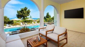 A balcony with a pool view, white cushioned chairs, a wooden table, and a black TV screen. - Sunset View Paradisi Villa (Rhodes)
