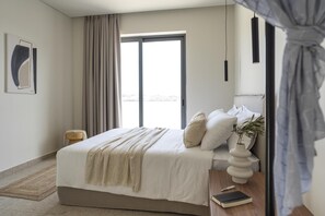 A modern bedroom with a large bed, a bedside table with a book and a lamp, a window with curtains, and a painting on the wall.