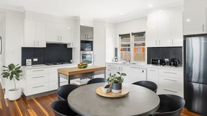 A modern kitchen with white cabinets, a central island, and a dining area with black chairs. - Blue View Cottage! Simply Perfect! (Centenary Heights)