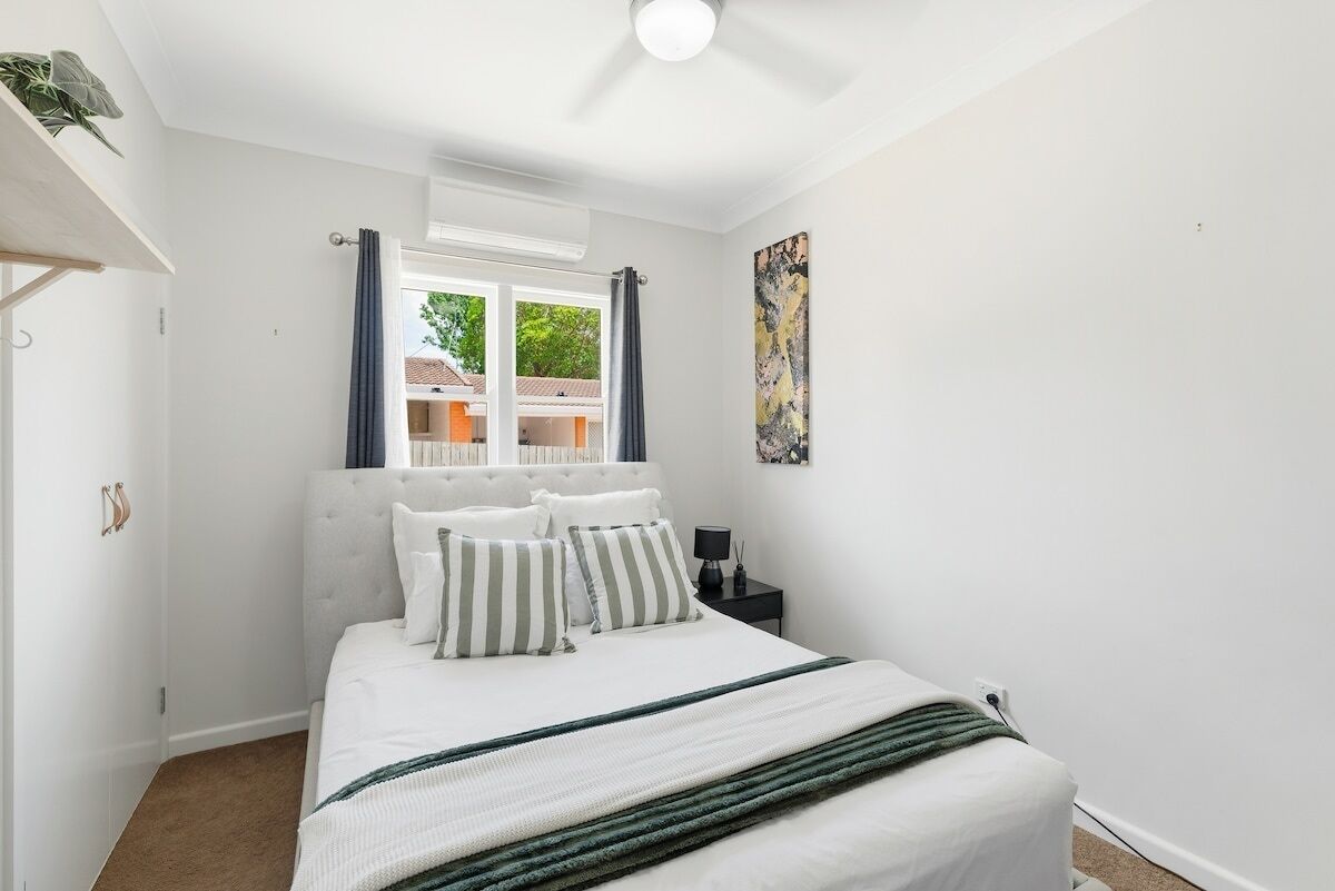 Una camera da letto con un letto, un comodino, una finestra con tende, una ventola a soffitto e un quadro appeso al muro.