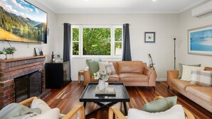 A living room with a fireplace, leather sofas, a coffee table, and a large window with curtains. - Blue View Cottage! Simply Perfect! (Centenary Heights)