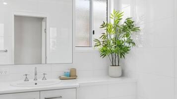 Bagno con lavandino bianco, due rubinetti, uno specchio, una finestra con vetro satinato e una pianta in vaso.