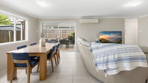 A modern living room with a wooden dining table, blue chairs, a beige sofa with a striped blanket, and a large window with a view of a fence and trees. - Middle Ridge Retreat – Walk to Shops & Parks! (Middle Ridge)