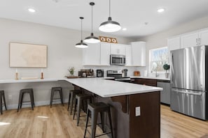 A modern kitchen with white cabinets, stainless steel appliances, a large island with bar stools, pendant lighting, and a view of the outdoors.