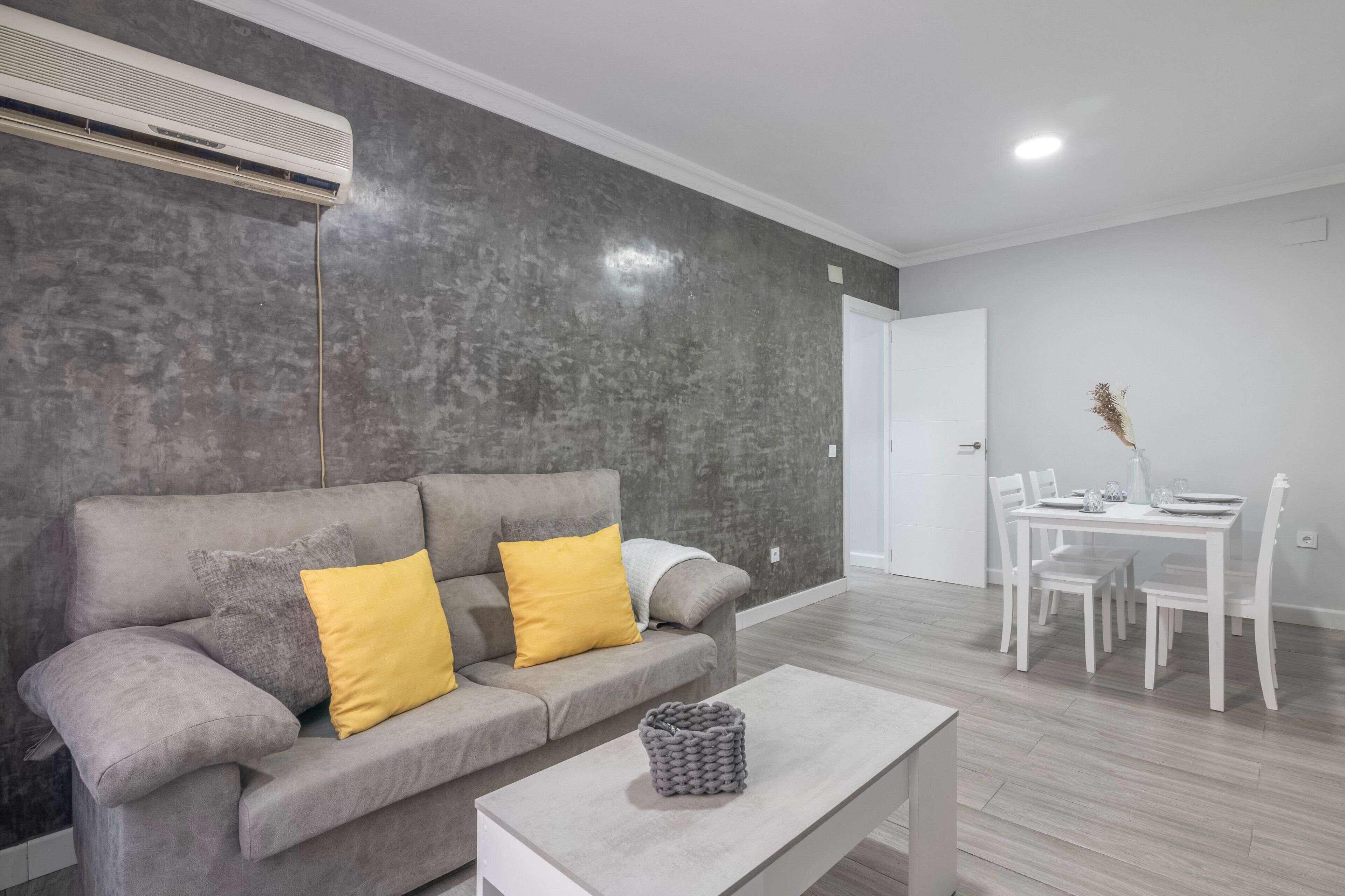 A modern living room with a grey sofa, two yellow cushions, a grey woven basket on a white coffee table, a dining area with a white table and chairs, and a grey textured wall.