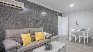 A modern living room with a grey sofa, two yellow cushions, a grey woven basket on a white coffee table, a dining area with a white table and chairs, and a grey textured wall.