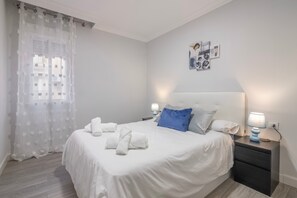 A neatly made bed with white linens and blue pillows, a bedside table with a lamp, and a window with sheer curtains. - Grey House Apartment (Huelva)