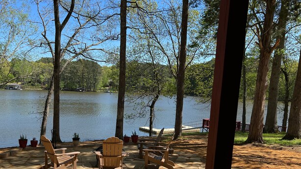 An outdoor seating area with wooden chairs and tables, overlooking a calm lake with trees in the background. - Cottage on Cypress Bay Near Paris Landing State Park (Buchanan)