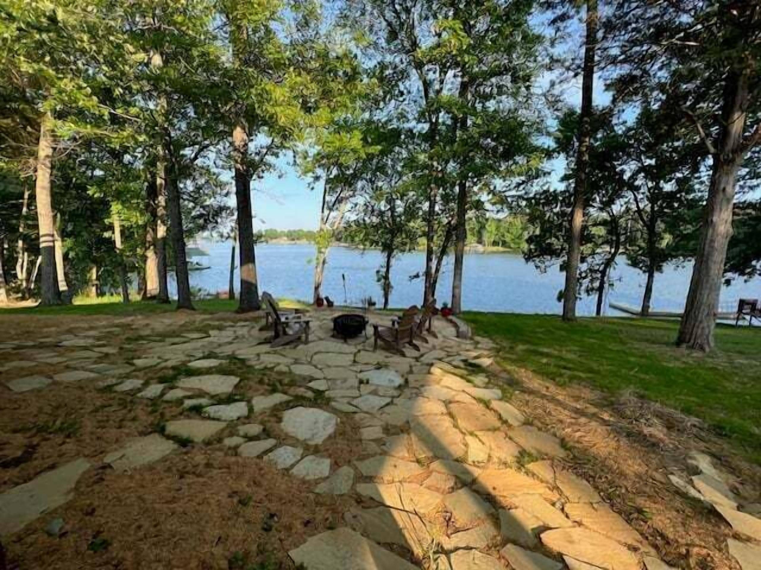 A stone-paved area with a fire pit, surrounded by trees and a lake view.