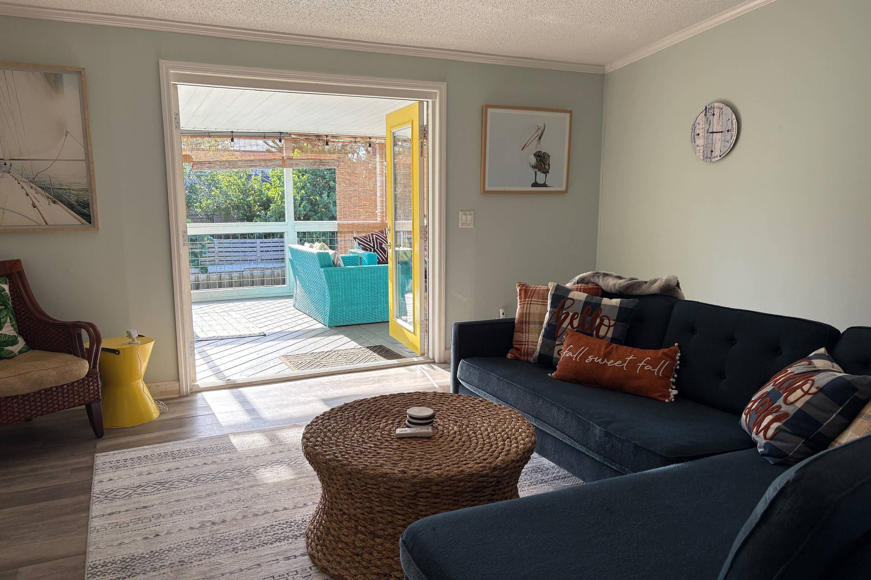 A living room with a blue sofa, a wicker ottoman, and a yellow side table. There is a large window with a view of a patio area.