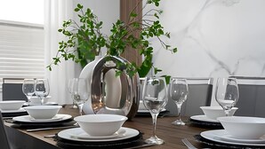 Een eettafel gedekt met witte borden, kommen en wijnglazen, omringd door een potplant.