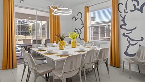 Un coin repas avec une table blanche dressée pour six personnes, entourée de chaises grises, agrémenté de rideaux jaunes et orné d’une grande fresque murale.