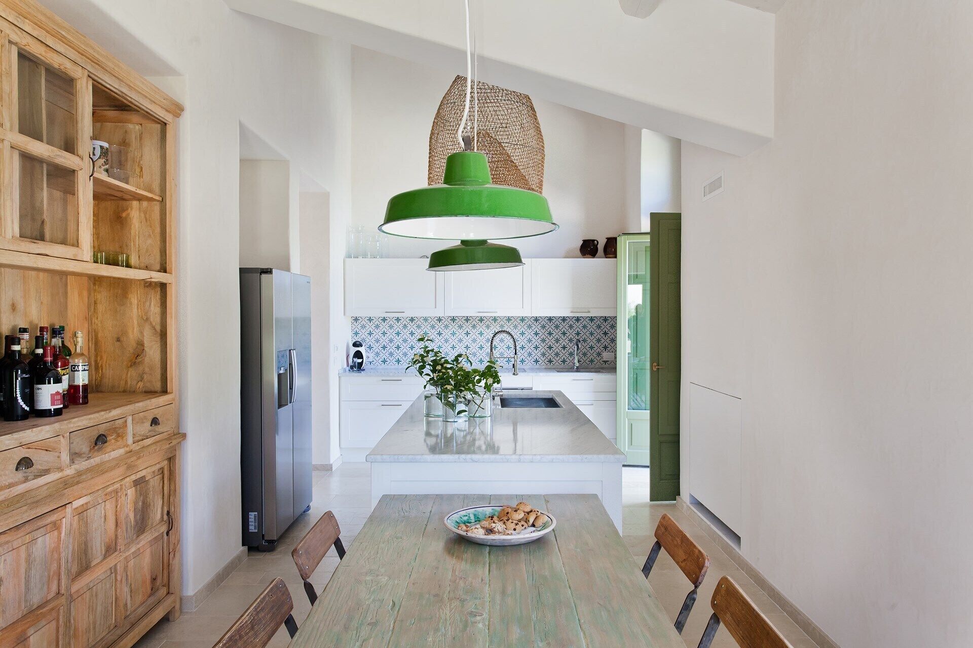 A modern kitchen with a wooden cabinet, a stainless steel refrigerator, a green pendant light, and a dining table with chairs.