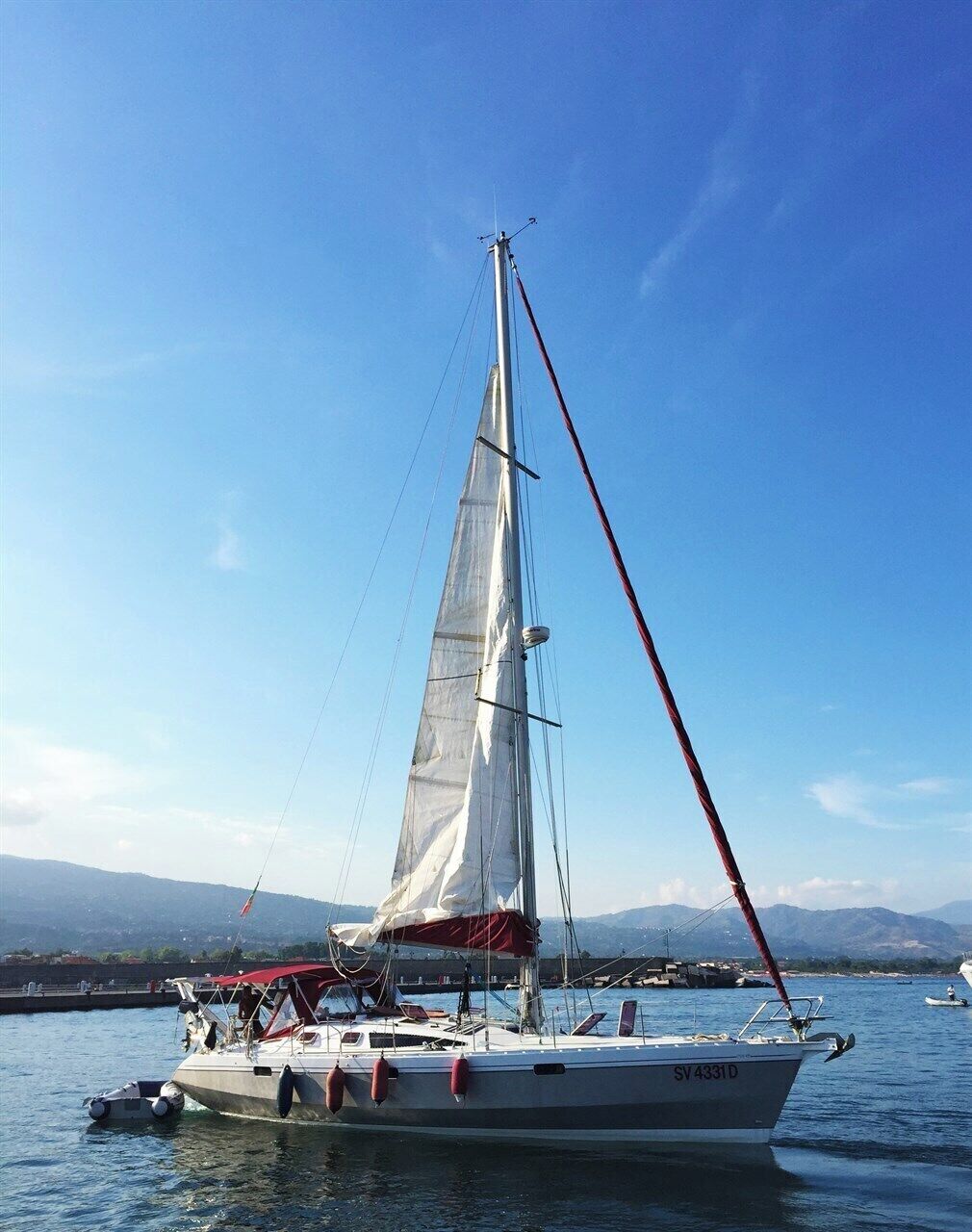 A sailboat with a white hull and red accents, docked at a marina with mountains in the background.