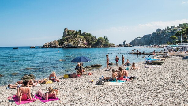 Een kiezelstrand met mensen die zonnebaden en zwemmen, een rotsachtige landtong in de achtergrond en een helderblauwe lucht.
