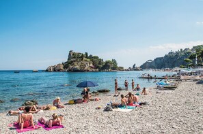 A pebble beach with people sunbathing and swimming, a rocky outcrop in the background, and a clear blue sky.