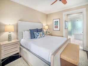 Une chambre d’hôtel avec un grand lit, deux tables de chevet, une petite table d’appoint, un ventilateur de plafond, une salle de bain visible à travers une porte ouverte et une œuvre d’art encadrée accrochée au mur.