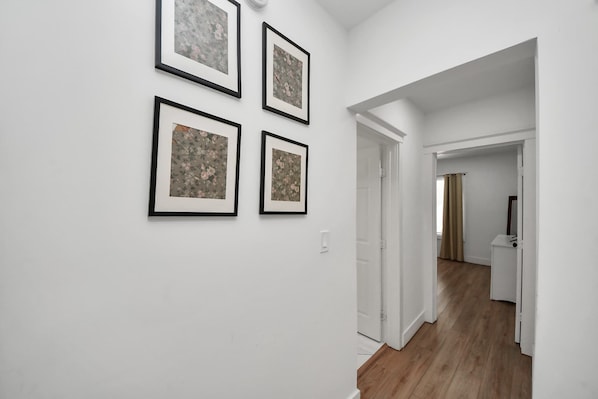 A hallway with white walls and wooden flooring, leading to a room with a door and a window.