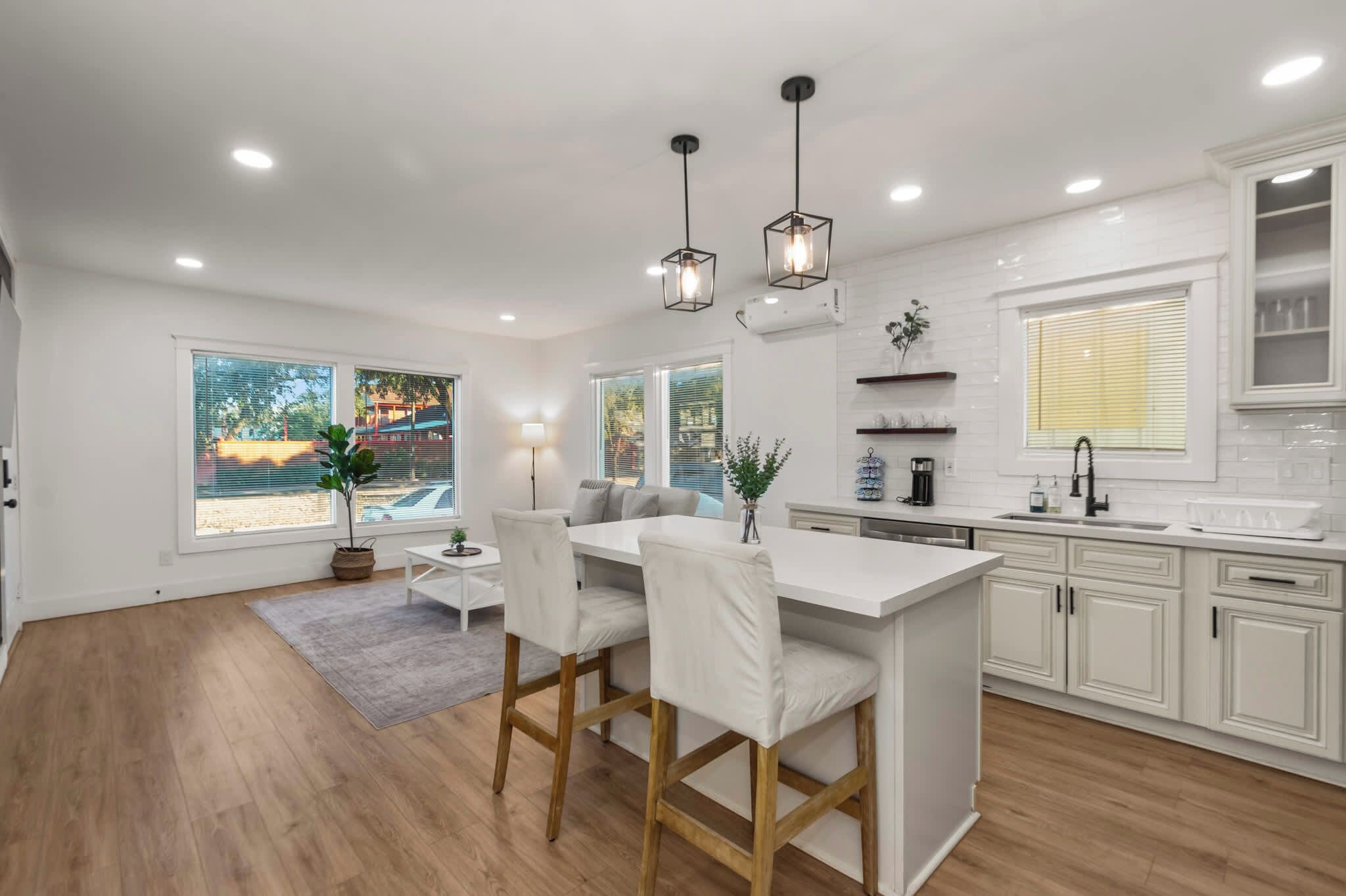 A modern kitchen with white cabinetry, a central island with bar stools, pendant lighting, and a view of the outdoors through large windows.
