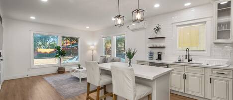 A modern kitchen with white cabinetry, a central island with bar stools, pendant lighting, and a view of the outdoors through large windows.