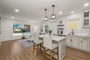 A modern kitchen with white cabinetry, a central island with bar stools, pendant lighting, and a view of the outdoors through large windows.