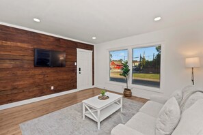 A modern living room with a flat-screen TV mounted on a wooden wall, a white sofa, a coffee table with a plant, and a large window offering a view of the outside.