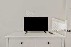 Un bureau blanc sur lequel est posée une télévision à écran plat, une télécommande et une porte fermée avec des stores.