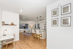 A modern kitchen with white cabinetry, a dining area with a white table and chairs, and a hallway with a white wall and framed pictures.