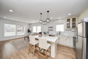 A modern kitchen with a central island, wooden chairs, pendant lighting, and white cabinetry.