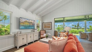 Un vaste salon agrémenté d’un grand canapé orange, d’un téléviseur à écran plat fixé au mur, d’un meuble en bois et offrant une vue sur un paysage tropical grâce à de grandes fenêtres.