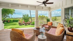 Une terrasse extérieure couverte, aménagée avec des meubles en osier, un ventilateur de plafond et offrant une vue sur une pelouse bien entretenue et des arbres.