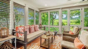 A spacious room with a large window offering a view of lush greenery. There is a sofa with orange cushions, a wicker chair, and a small table with decorative items. - Resort Amenities | Golf Course View | Cottage + Guesthouse (Koloa)
