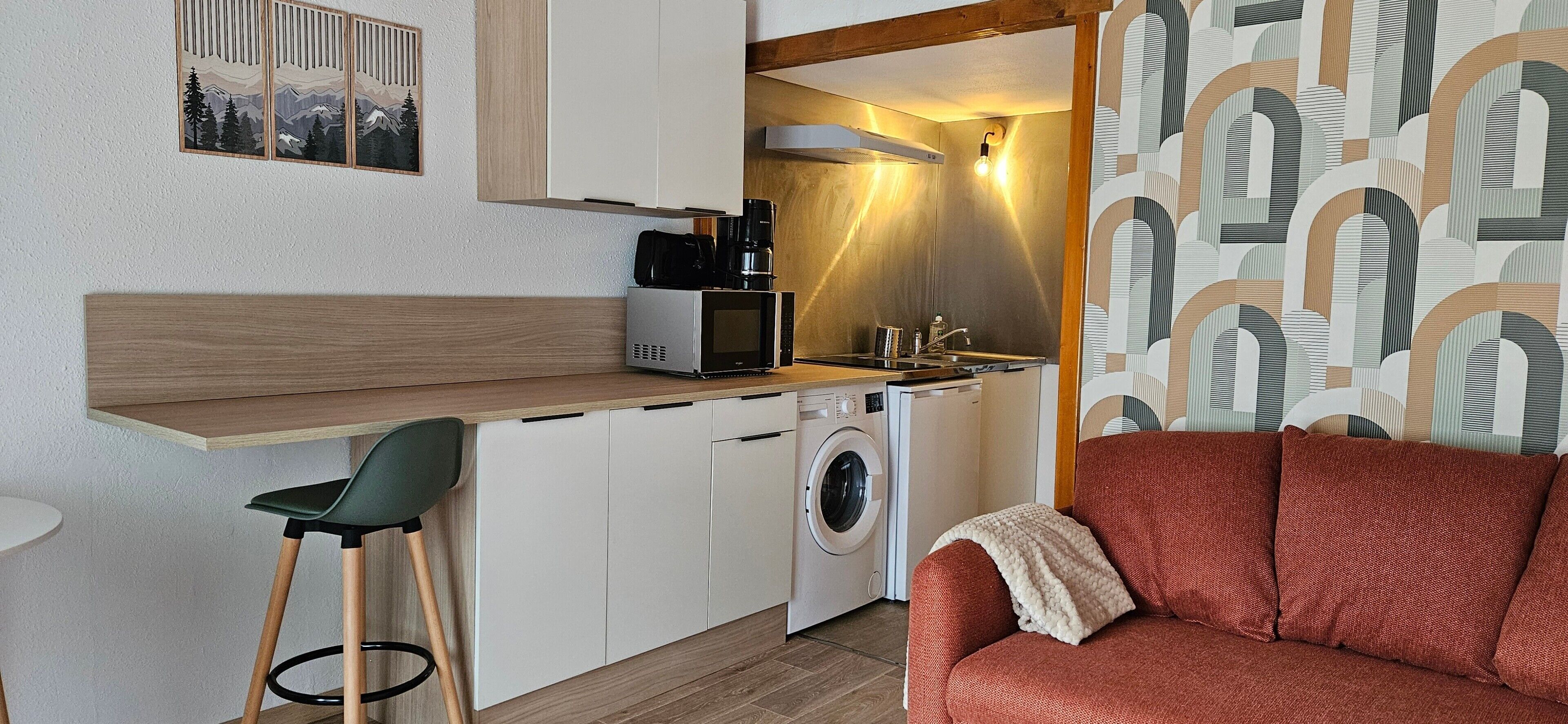 Eine kompakte Küche mit Mikrowelle, Kaffeemaschine und Waschmaschine, ein kleiner Essbereich mit einem Stuhl und ein rotes Sofa in modernem Ambiente.