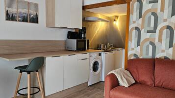 Eine kompakte Küche mit Mikrowelle, Kaffeemaschine und Waschmaschine, ein kleiner Essbereich mit einem Stuhl und ein rotes Sofa in modernem Ambiente.